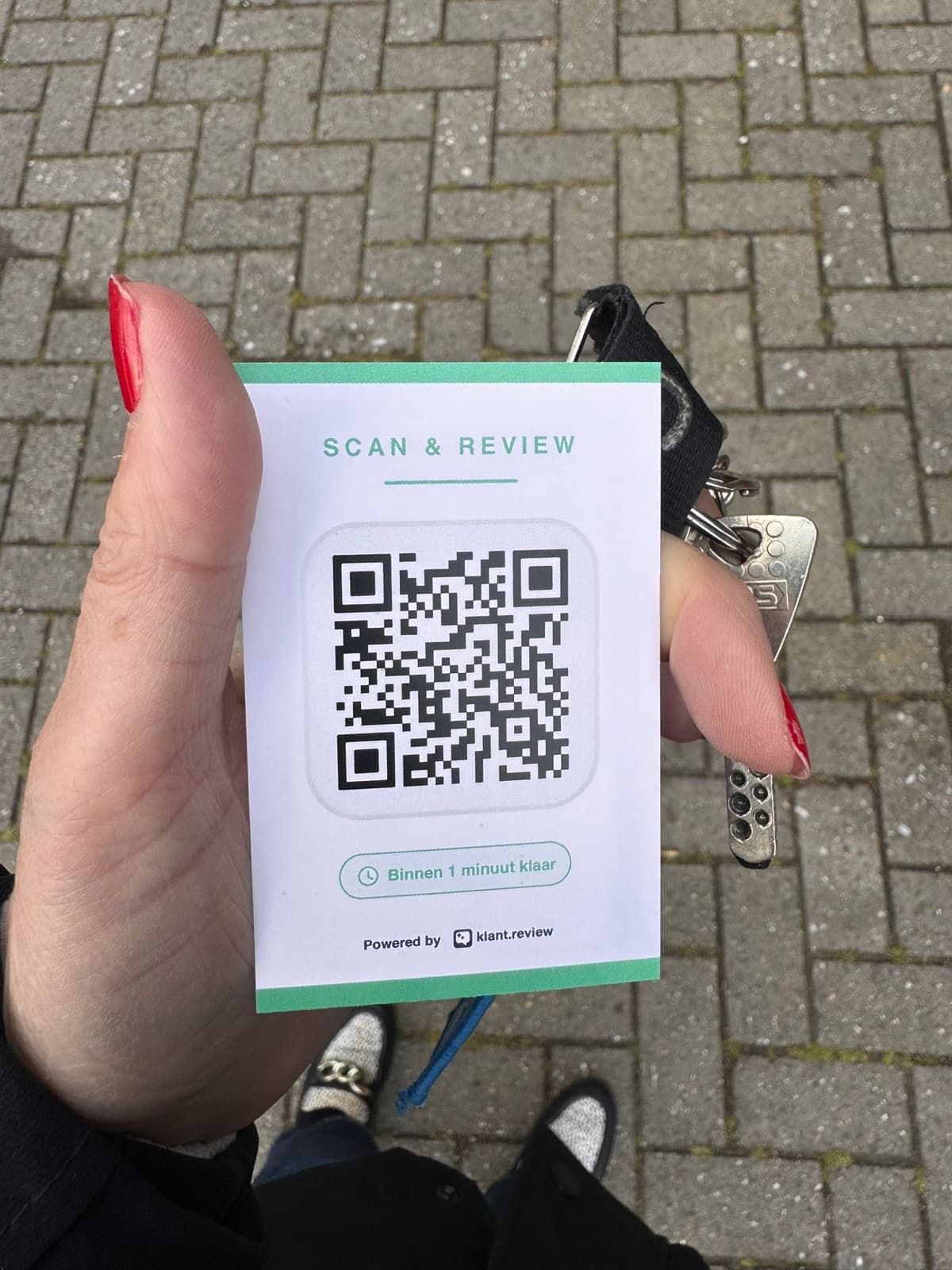 Voorbeeld-QR-review-kaartje met 'Scan & Review' en 'Binnen 1 minuut klaar', vastgehouden naast sleutels op straat